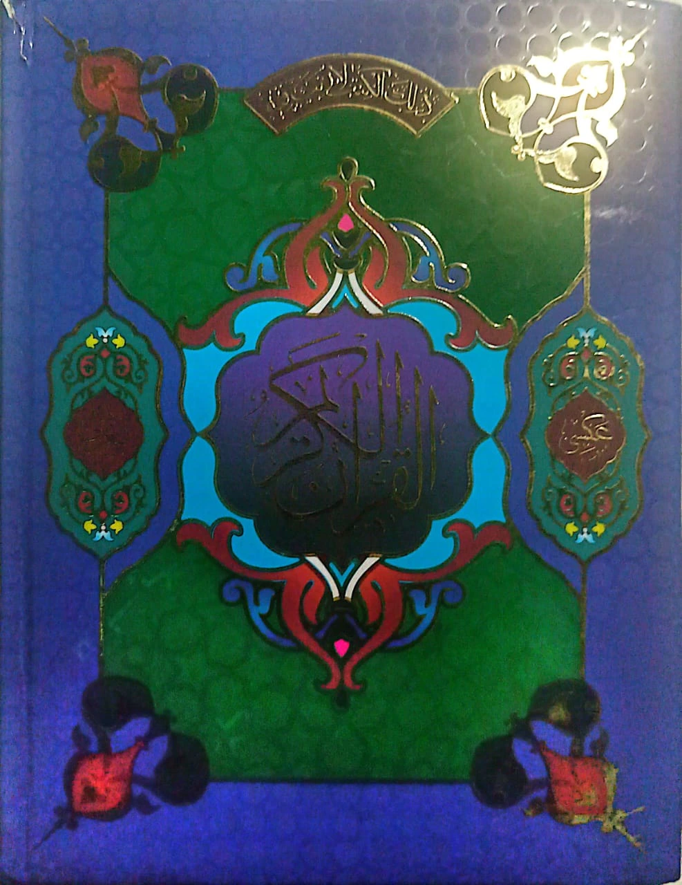 A decorative gift edition of the Quran Majeed in Arabic, featuring a colorful cover with intricate blue, green, and red patterns, central Arabic calligraphy, and gold detailing. Product: আরবি কুরআন মাজিদ – রঙিন ডিজাইন কভার.